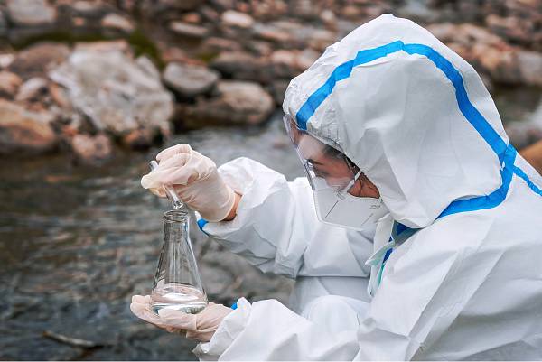 Person in Schutzanzug entnimmt Wasserprobe aus einem Fluss zur Analyse möglicher Schadstoffbelastung.