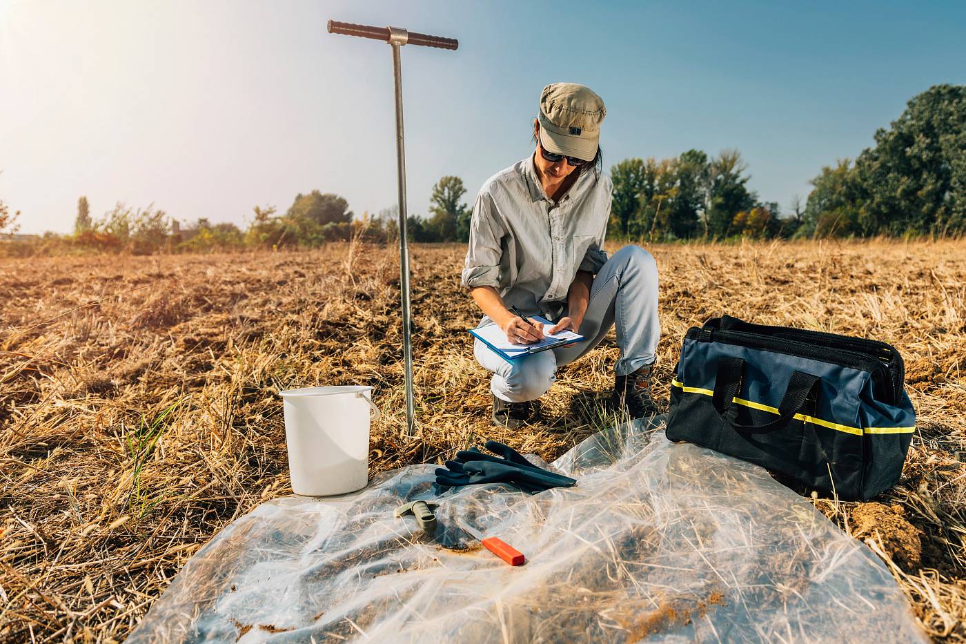 Geoökologe führt Bodenuntersuchung auf einem Feld durch, notiert Ergebnisse auf einem Klemmbrett, umgeben von Probenequipment und Werkzeugen.