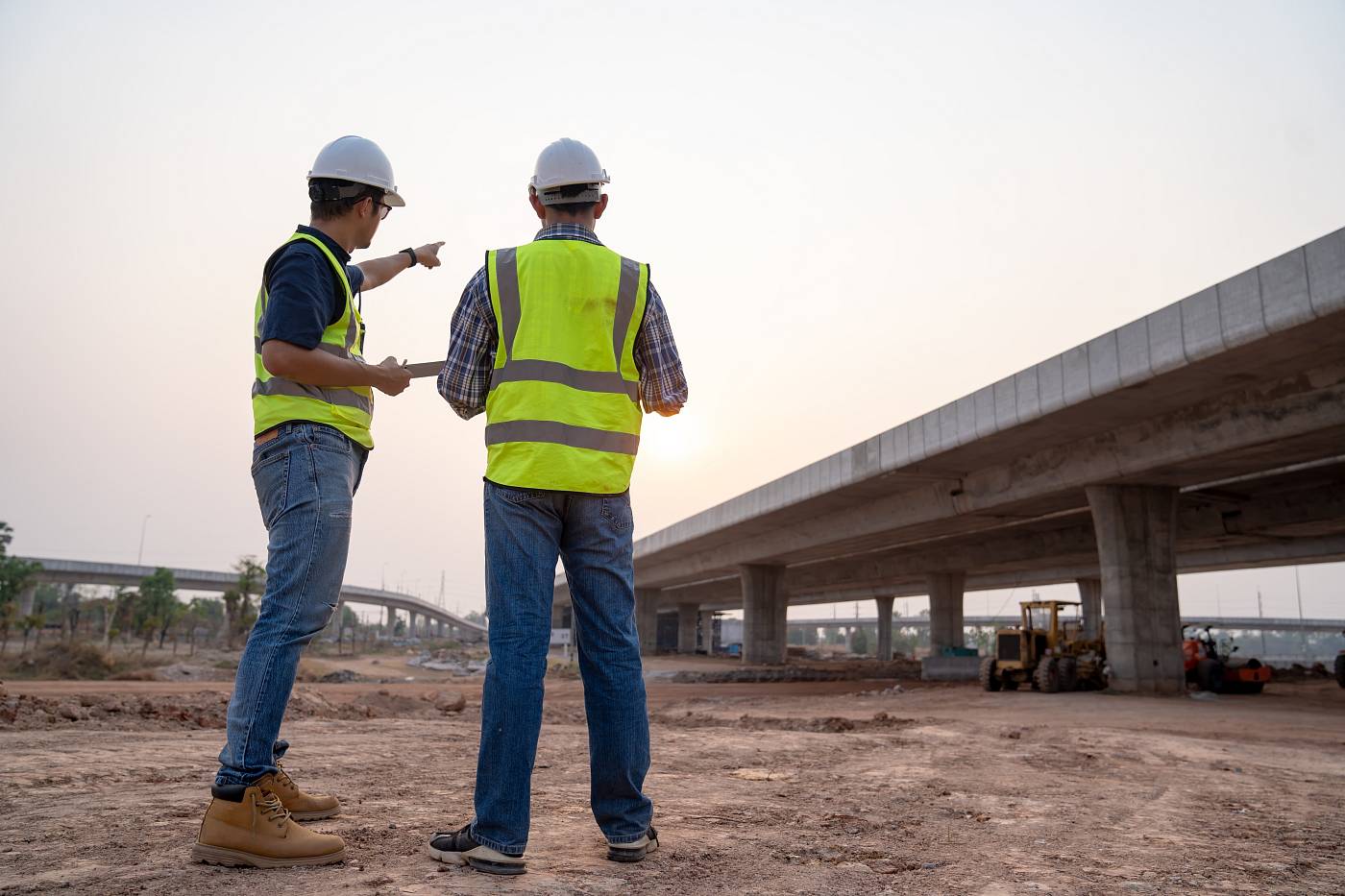 Zwei Bauingenieure in Warnwesten und Helmen begutachten eine Betonbrücke auf einer Baustelle und besprechen den Zustand der Baustruktur bei Sonnenuntergang.