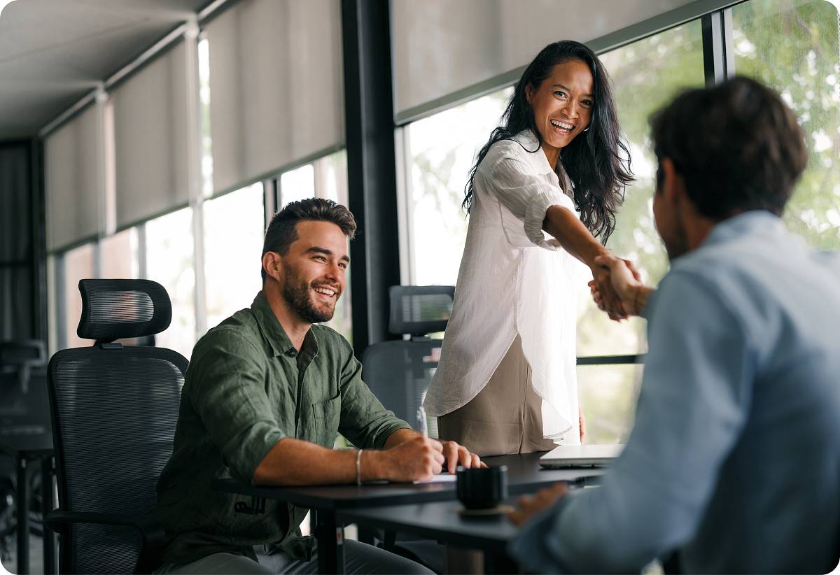 Geschäftliches Treffen: Frau begrüßt einen Mann per Handschlag, während ein Kollege lächelnd zusieht.