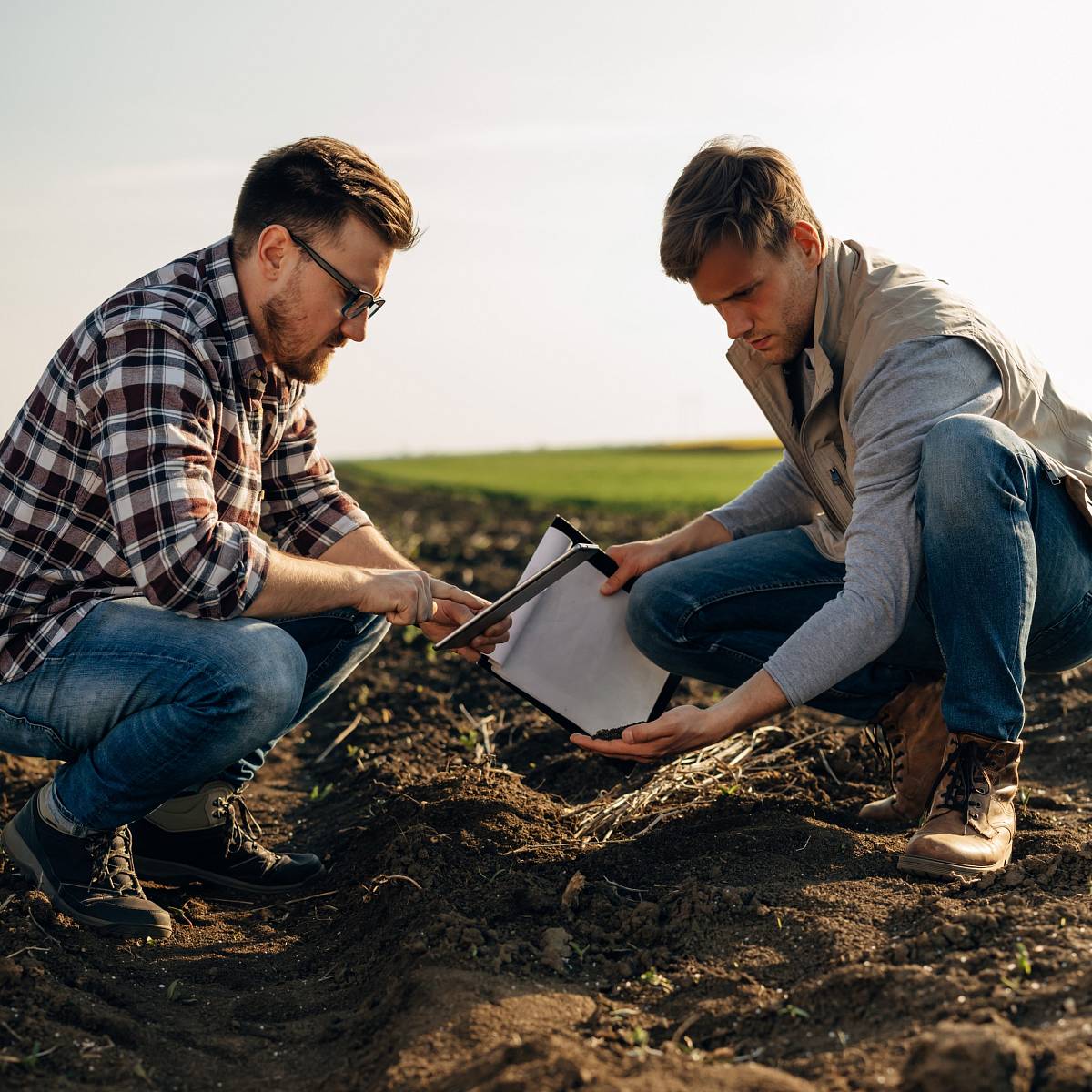 Zwei Fachleute untersuchen auf einem Feld Bodenproben und notieren Ergebnisse auf einem Klemmbrett.