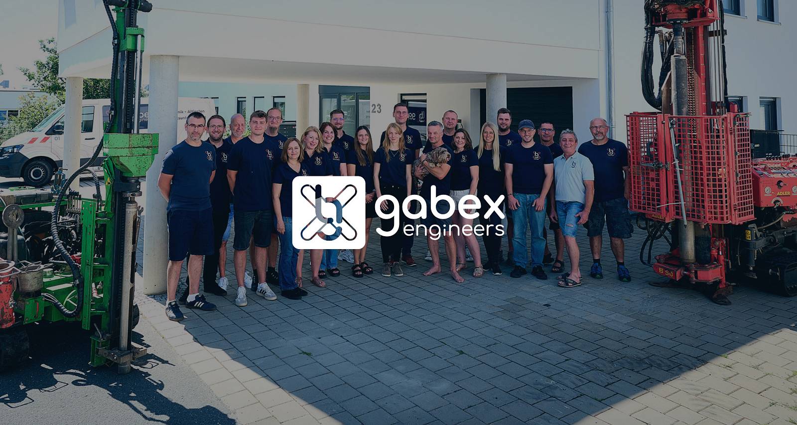 Team von gabex engineers vor dem Firmengebäude, gruppiert zwischen zwei großen Bohrgeräten in Grün und Rot, davor das Logo von gabex engineers.