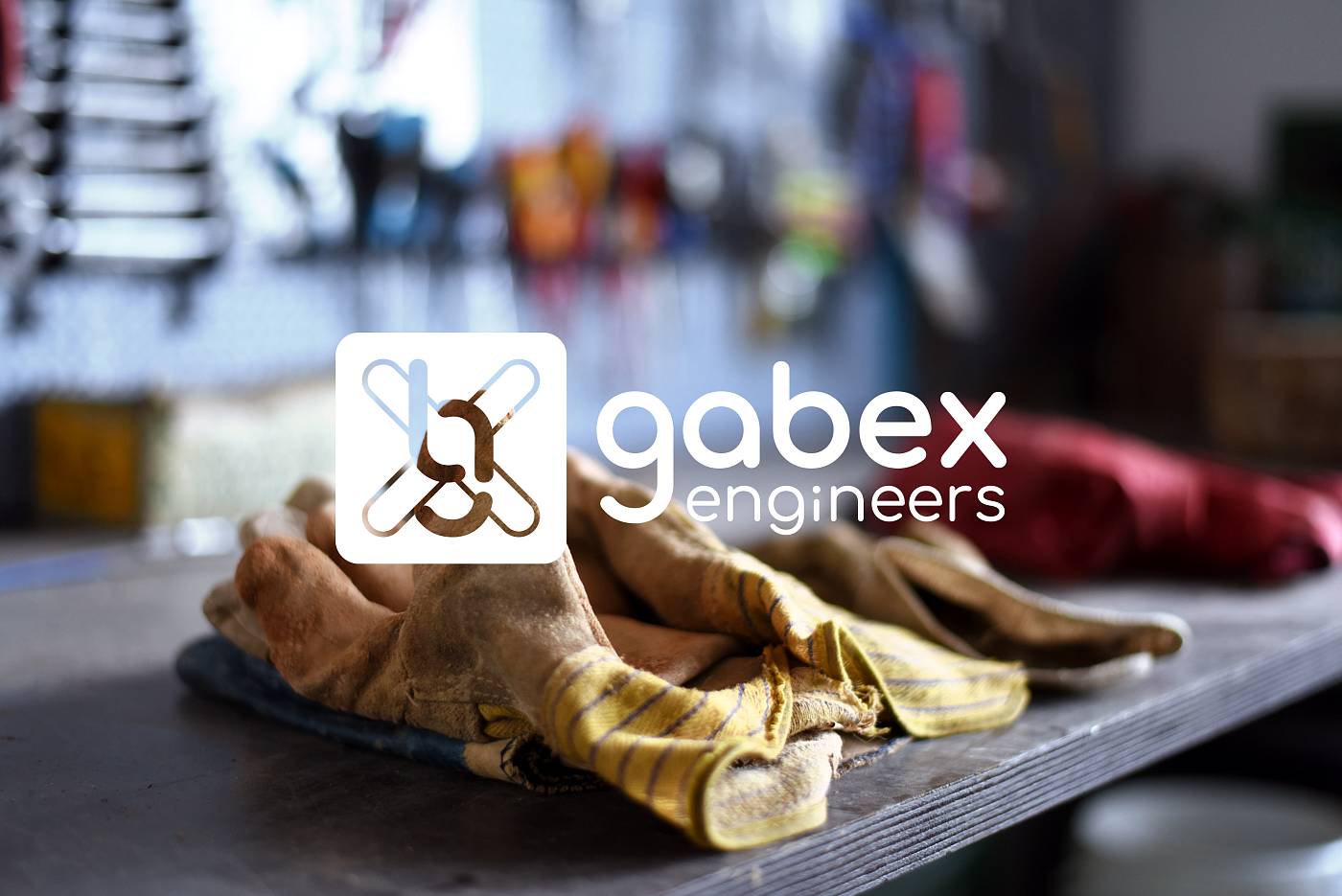 Arbeitshandschuhe auf einer Werkbank mit unscharfem Hintergrund und dem Logo von gabex engineers im Vordergrund.
