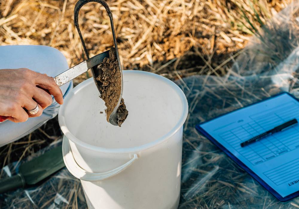 Bodenprobe wird mit einer Schaufel in einen weißen Eimer gefüllt, daneben liegt ein Klemmbrett mit Notizen.