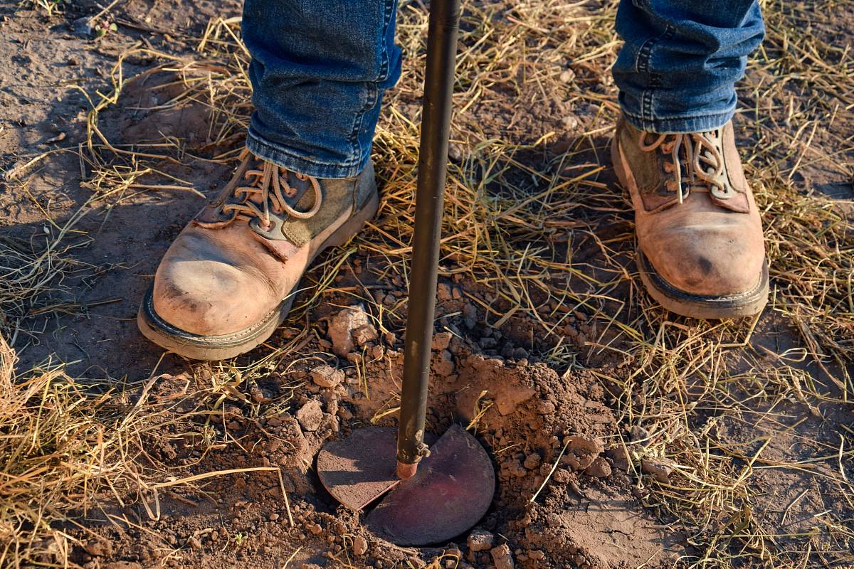 Nahaufnahme von Arbeitsschuhen und einer Erdsonde bei einer Bodenuntersuchung im Feld.
