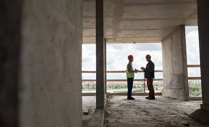 Zwei Bauingenieure mit Schutzhelmen und Warnwesten besprechen sich im Rohbau eines Gebäudes.