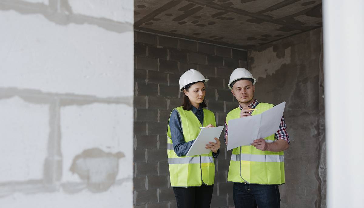 Zwei Fachleute mit Schutzhelmen und Warnwesten begutachten Baupläne und notieren Ergebnisse bei einer Baustellenbegehung.