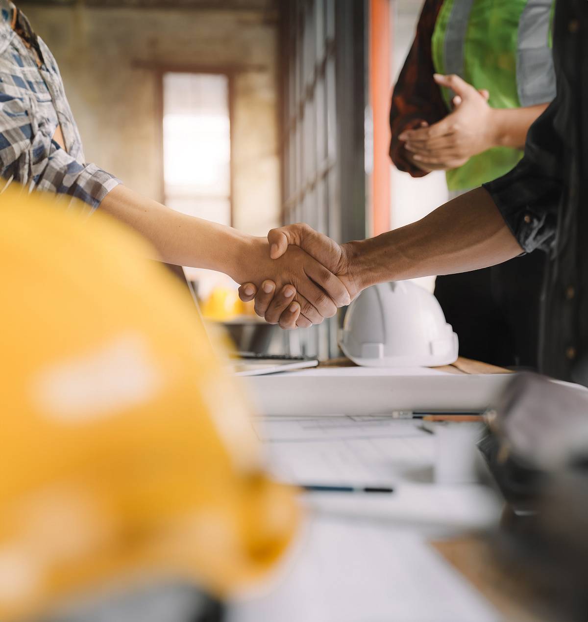 Zwei Personen geben sich auf einer Baustelle die Hand, im Vordergrund liegen Baupläne und Schutzhelme