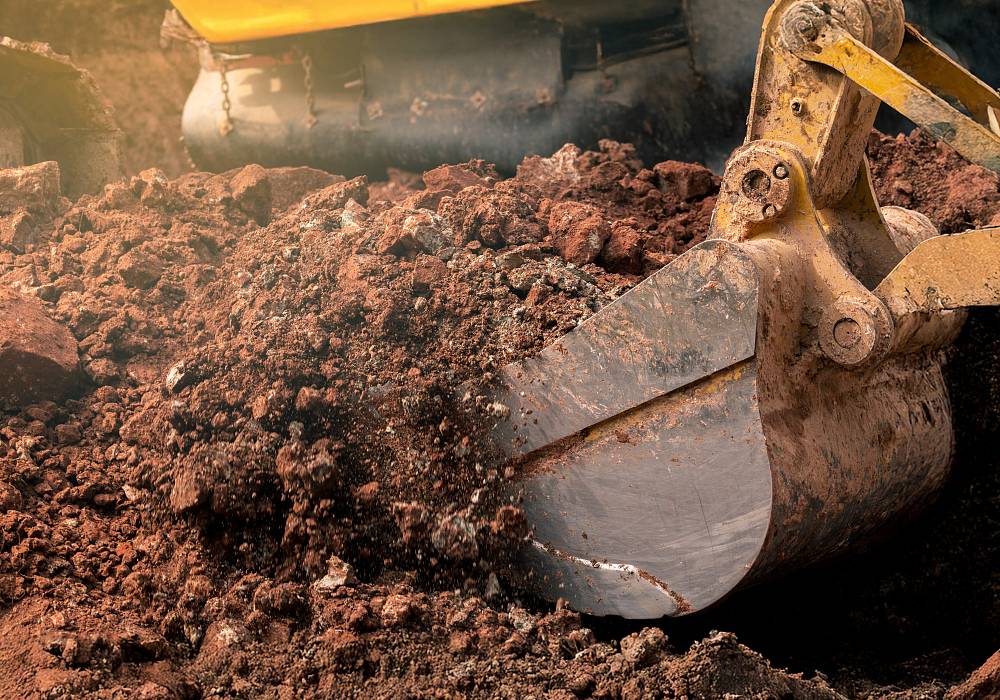 Baggerlöffel hebt rötlich-braune Erde aus.