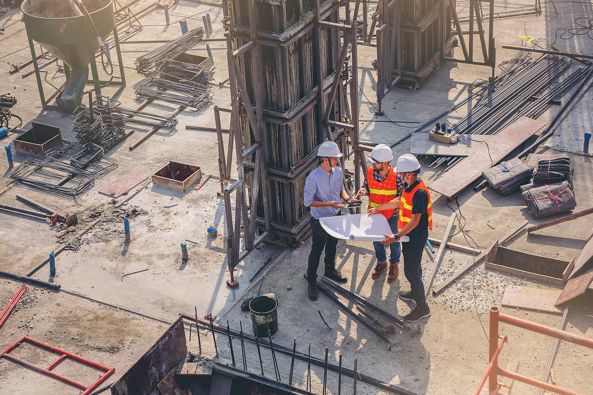 Drei Bauingenieure mit Schutzhelmen und Warnwesten besprechen Baupläne auf einer Baustelle.