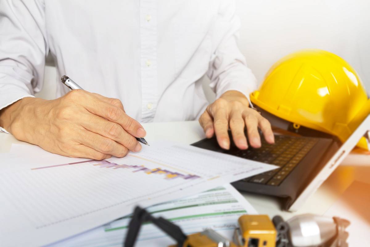 Ingenieur erstellt einen technischen Bericht am Schreibtisch, analysiert Bauunterlagen und trägt Daten in den Laptop ein – im Vordergrund liegen Diagramme und ein Sicherheitshelm.