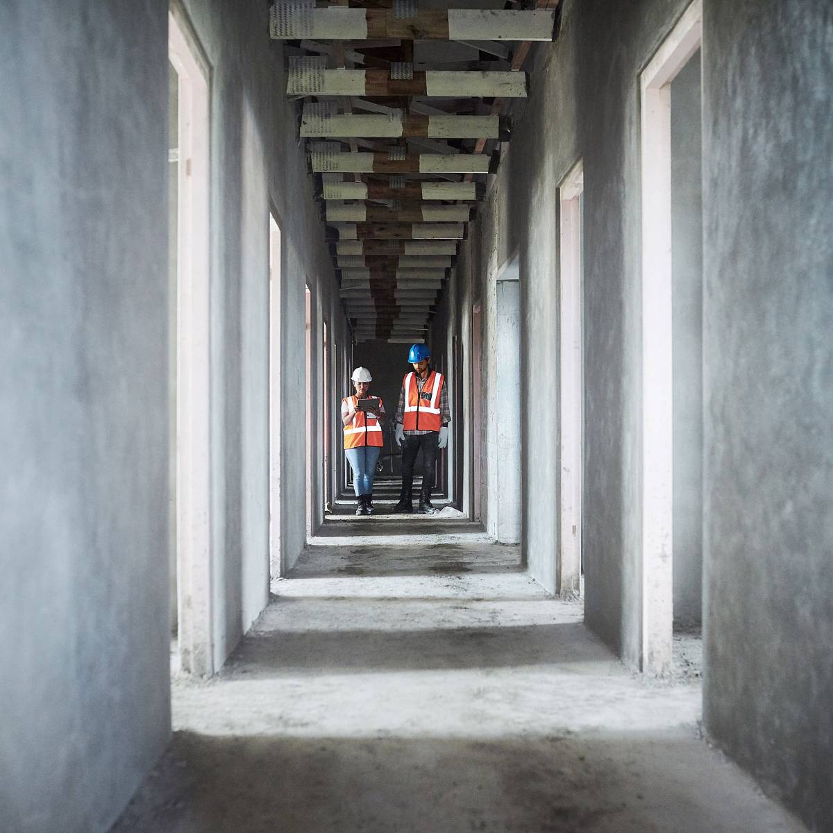 Zwei Ingenieure in Warnwesten und Schutzhelmen begehen einen unfertigen Betonbau und dokumentieren den Zustand des Gebäudes