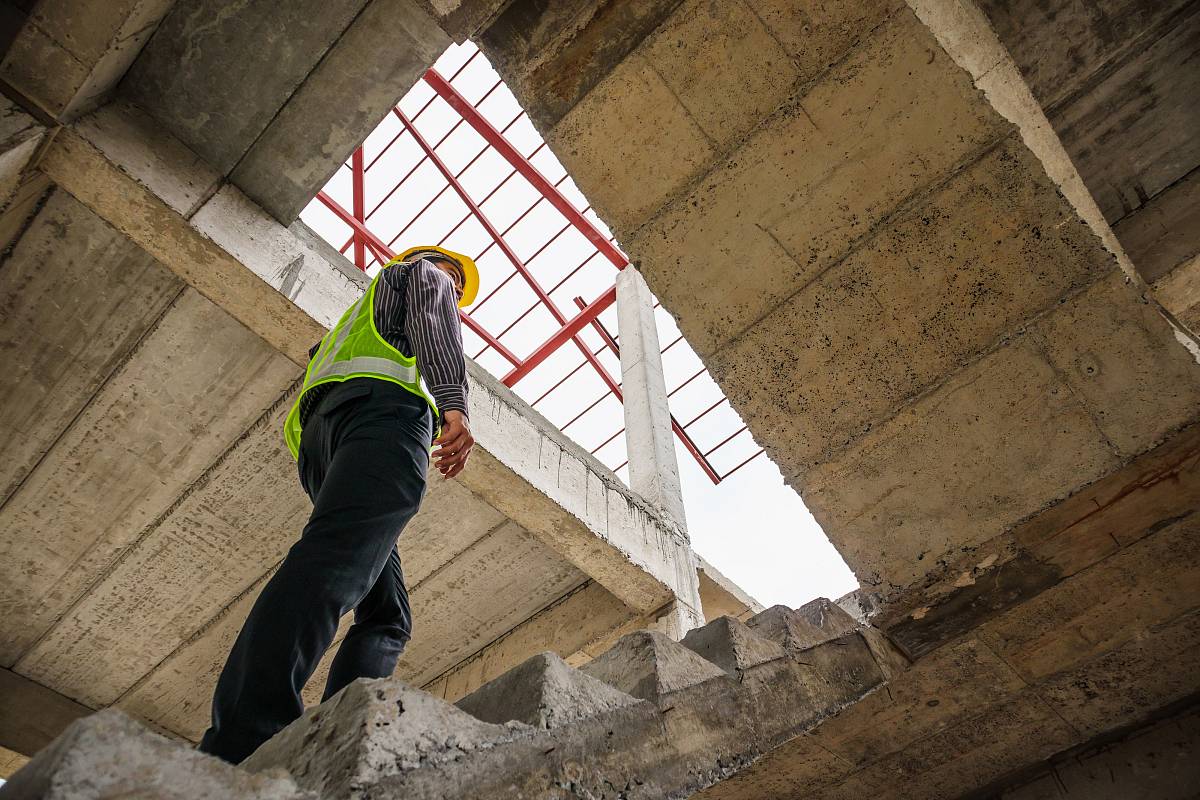 Ein Ingenieur mit Schutzweste und Helm begutachtet die Bausubstanz eines Rohbaus und steigt eine Betontreppe hinauf