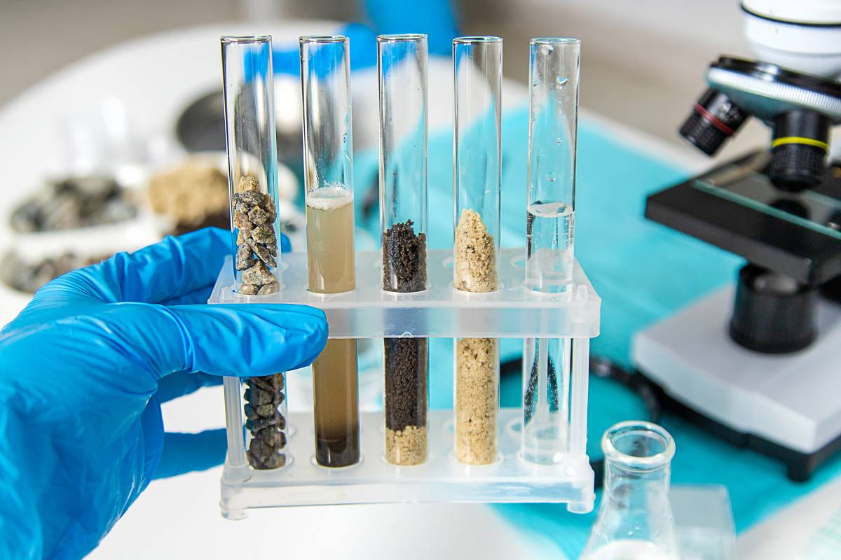 Nahaufnahme einer Hand mit blauen Schutzhandschuhen, die mehrere Reagenzgläser mit unterschiedlichen Boden- und Gesteinsproben hält.